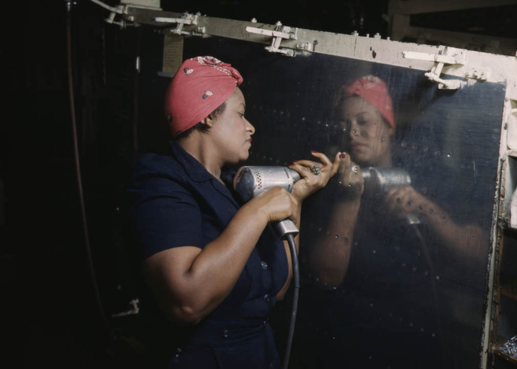 A woman, who is wearing the classic Rosie the Riveter outfit with a red scarf on her head and a blue jumpsuit, working on a Vengeance dive bomber airplane.