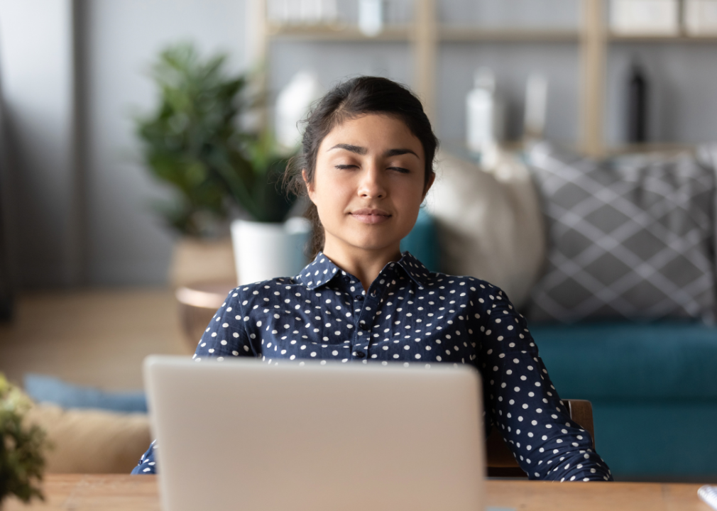 A calm-looking woman sitting with her eyes gently closed in front of a laptop in a home office.