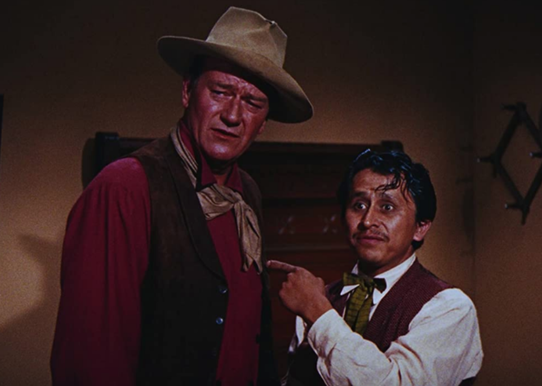 A man in a long sleeve shirt and vest points at John Wayne, in western wear.