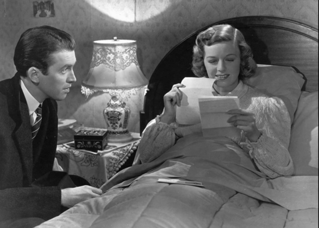 A woman reads a letter in bed while a man in a suit sits next to her bedside.