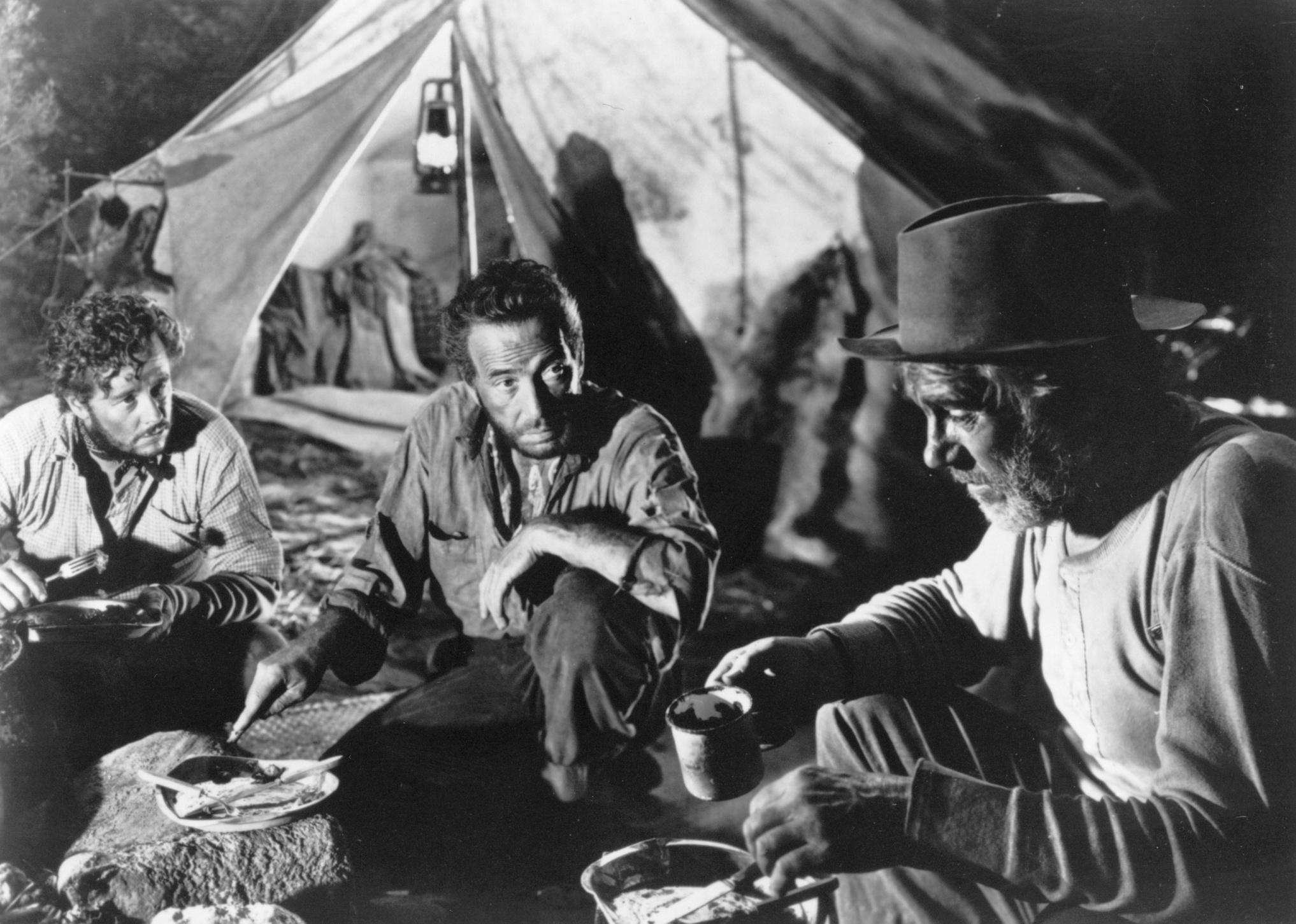 Humphrey Bogart and two other men eating in front of a tent.