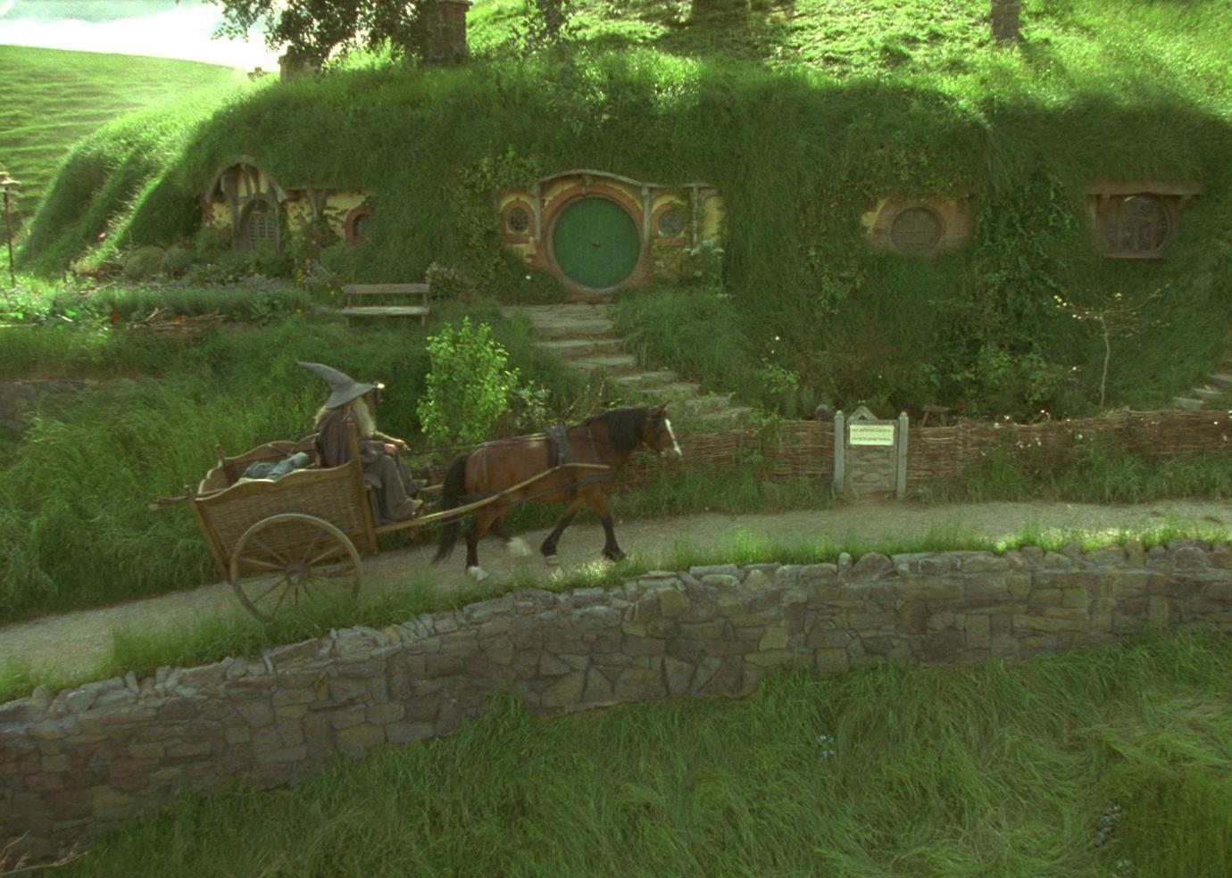 A wizard in gray with a pointy hat travels on a horse and cart up a green hill.