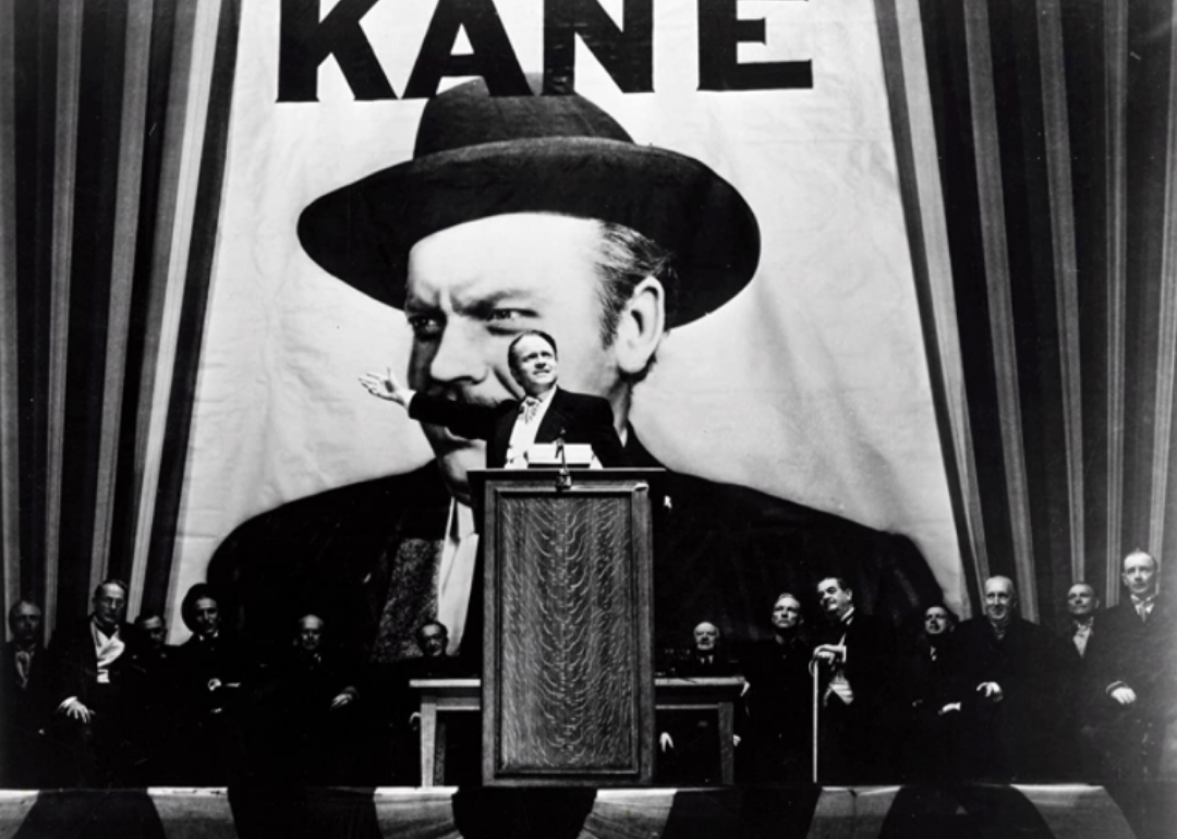 A man at a podium giving a speech in front of a large Kane sign.