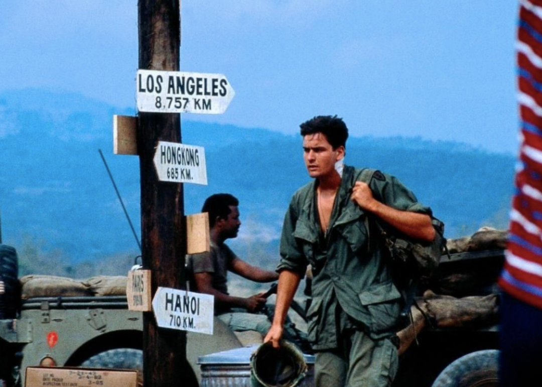 Charlie Sheen in military gear walking past a military jeep.