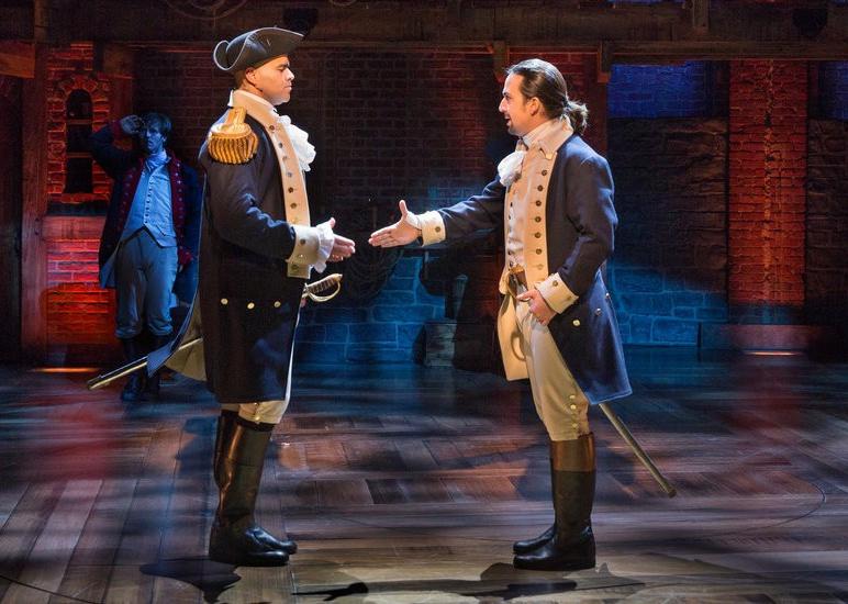Lin-Manuel Miranda and Chris Jackson shaking hands onstage dressed as founding fathers.