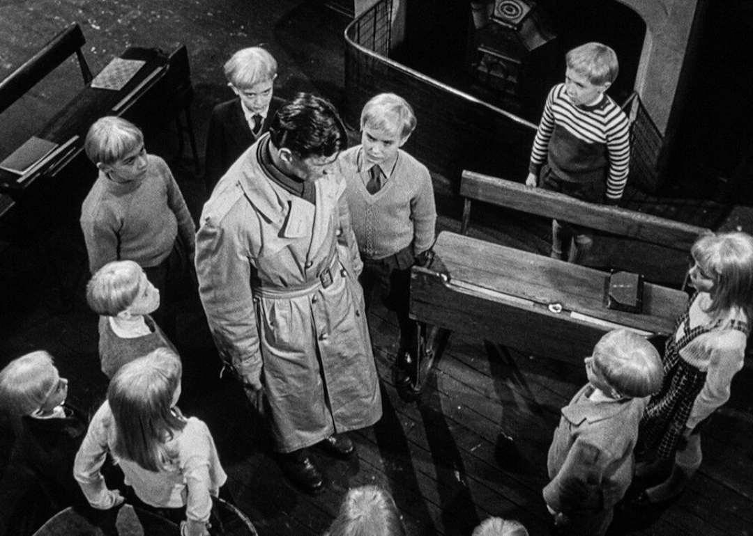 A man in a trenchcoat stands in the middle of a circle of children staring at him blankly.