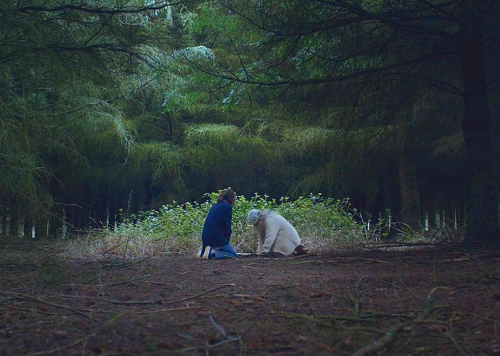 Two women crouched over something buried in the ground in the forest.