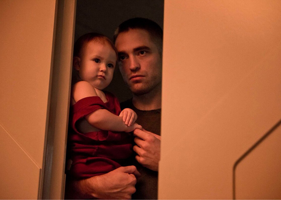 Robert Pattinson standing in a doorway holding a baby.