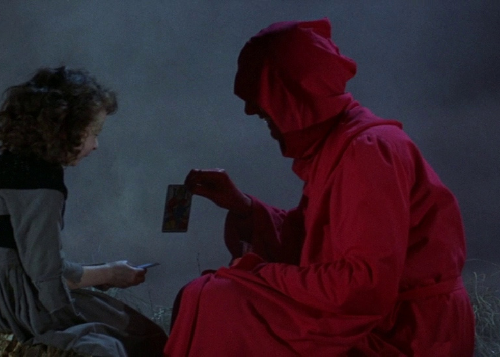 A person wearing a red hooded robe laughs while handing a child a card in the woods at night.