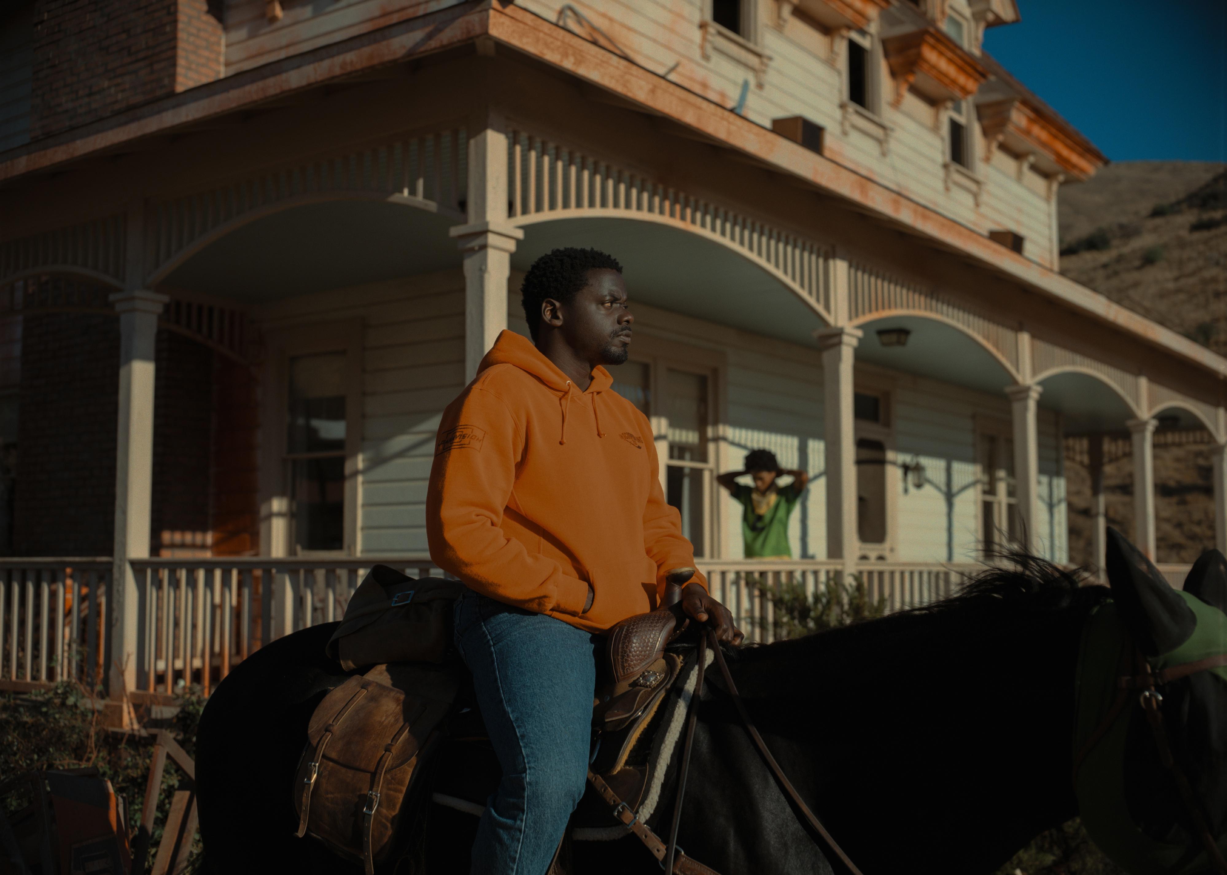 A man riding a horse in front of a large historic home with a woman on the porch.