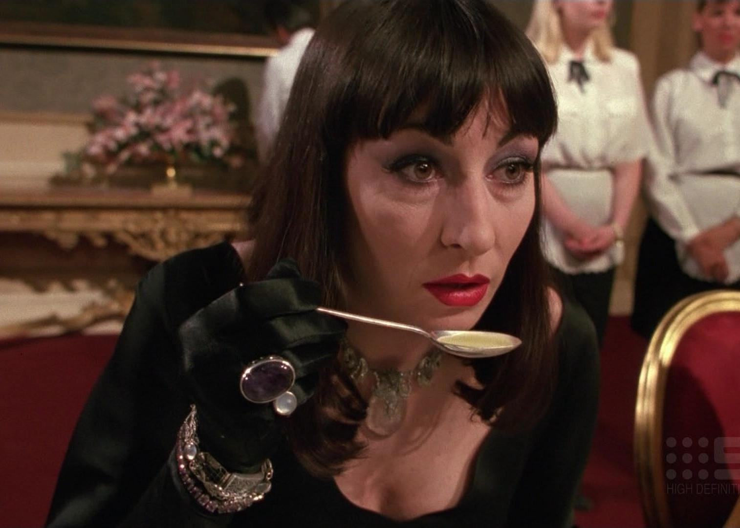 Anjelica Huston leaning in to taste from a spoon in a formal dining room.