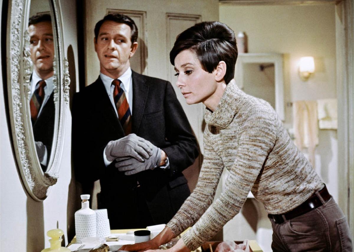 Audrey Hepburn stands in front of a dressing table and mirror while a man wearing gloves talks to her.