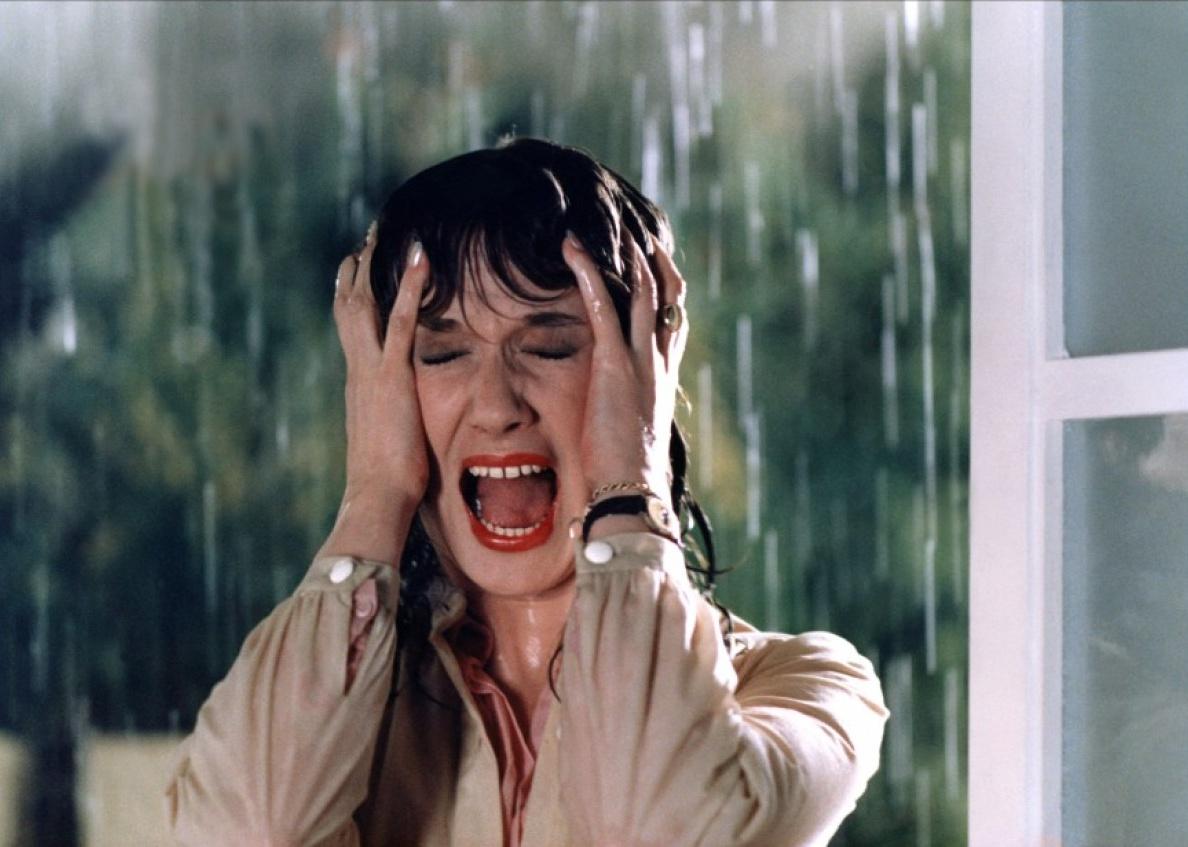 A woman soaking wet from the pouring rain screams clutching her head.
