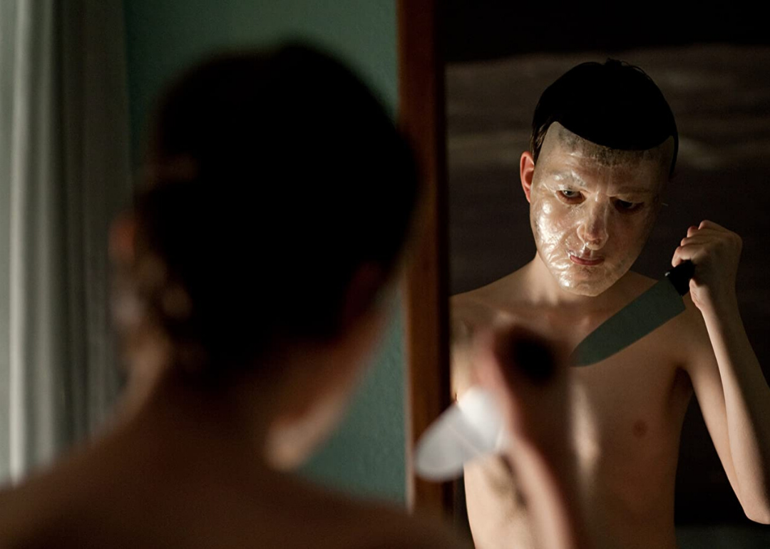 A dark haired man wearing a clear face mask standing in front of a mirror with a knife.