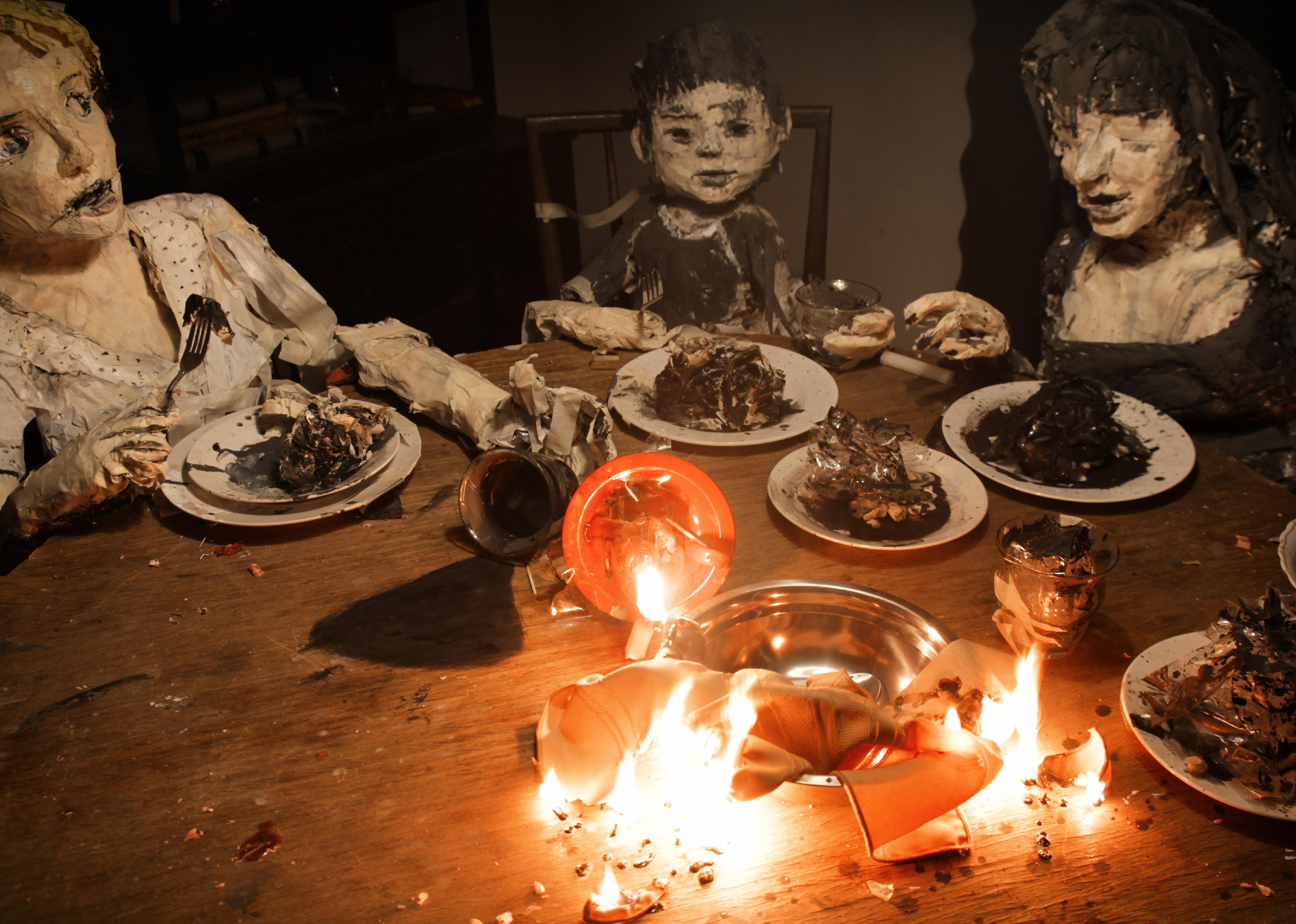 A family of paper dolls eats dinner at a table with burning bowls in the center.
