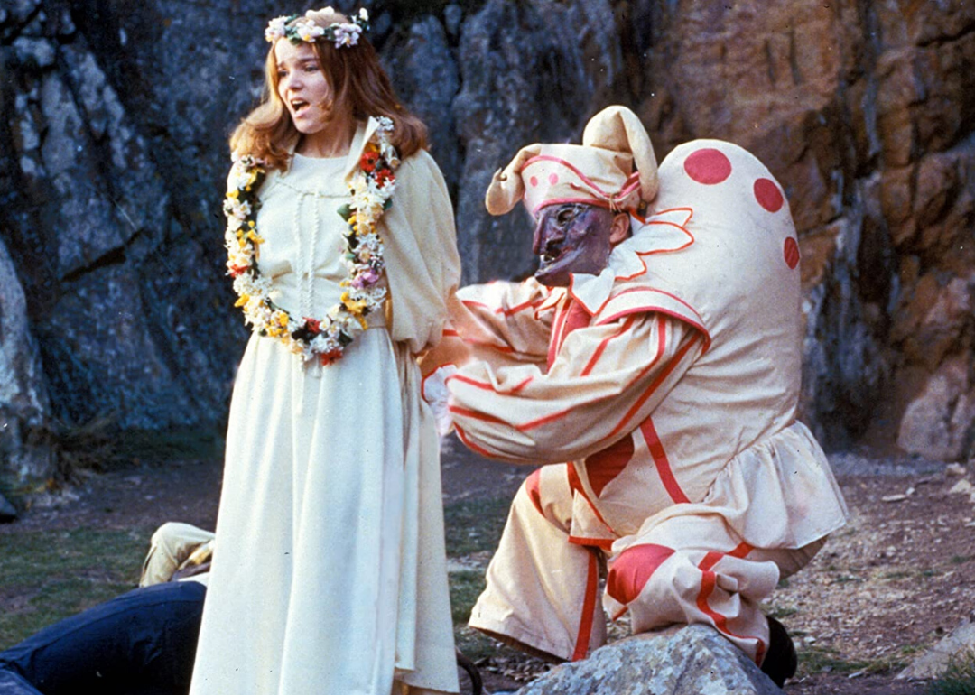 A woman dressed in white with a flower crown being tied up by a clown.