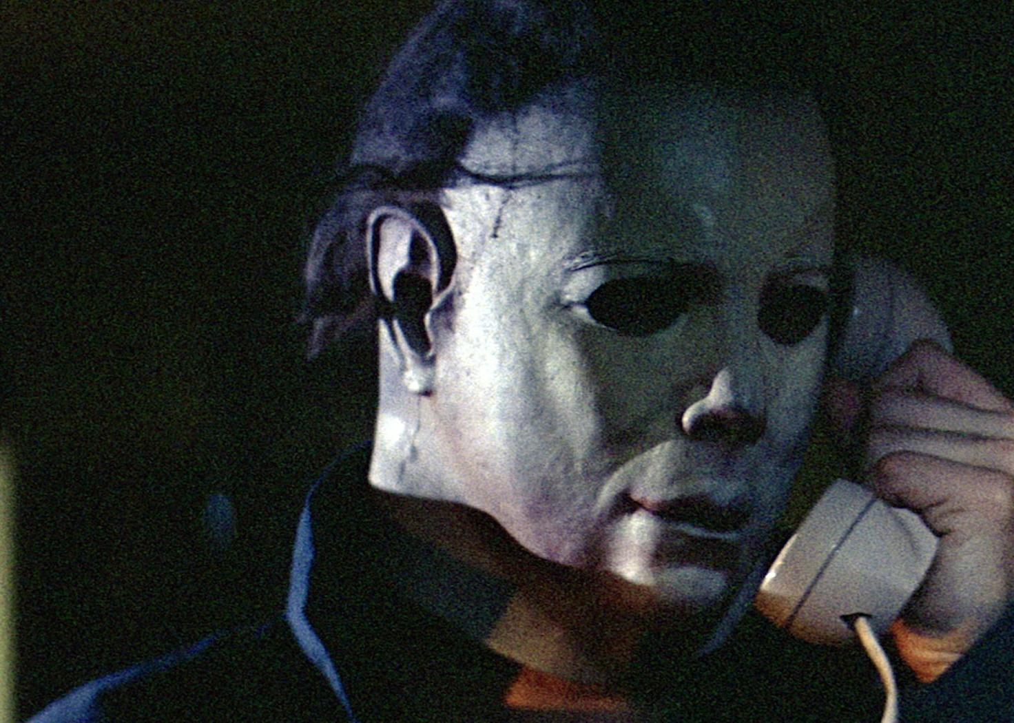 The killer, Michael Myers, wearing a white face mask and talking on the phone.