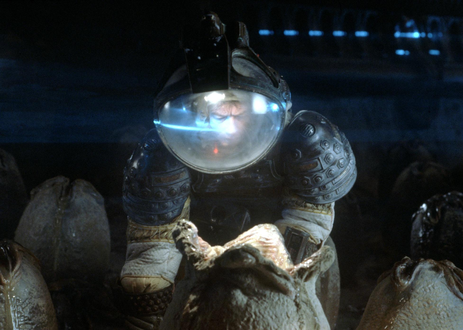 John Hurt, wearing a space suit and helmet, investigates living pods in the ground.