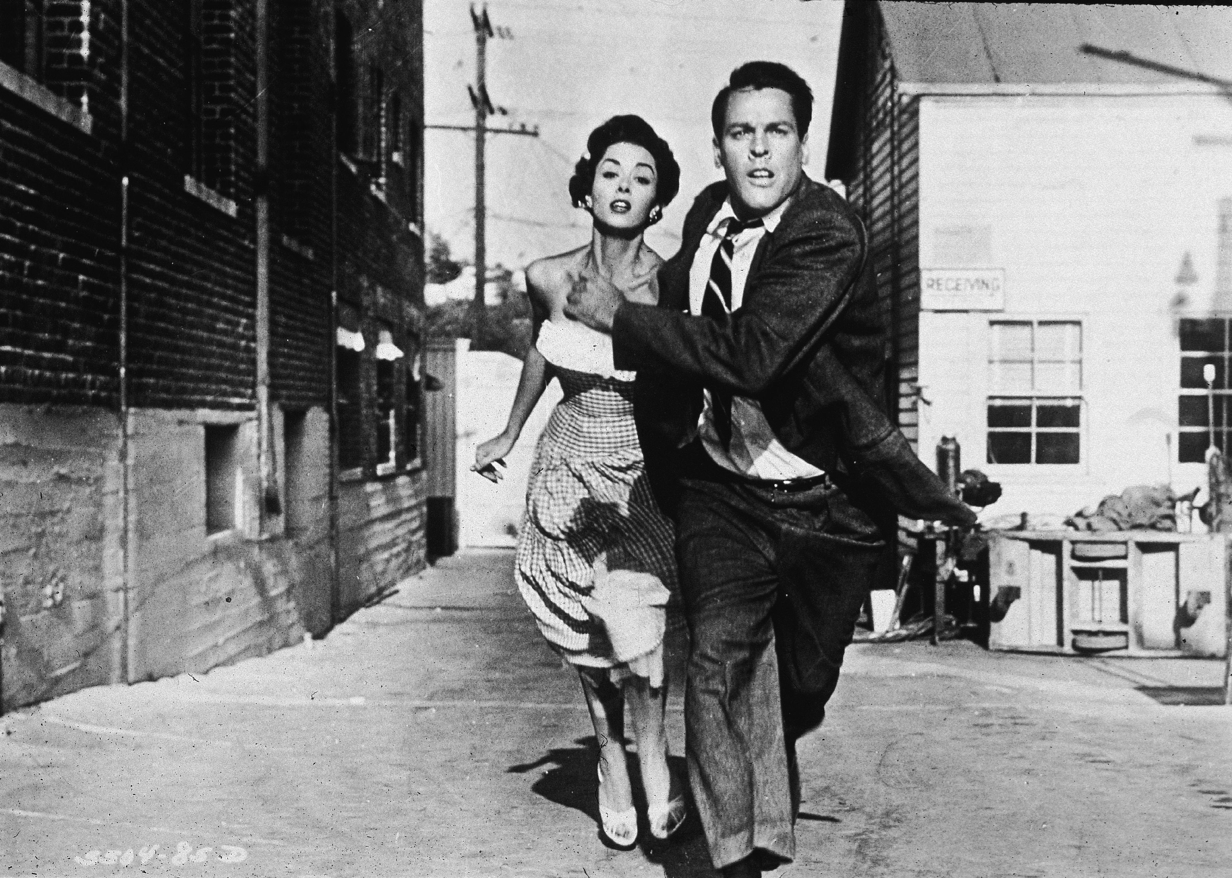 A man in a suit and a woman in a strapless dress run down the street looking scared.