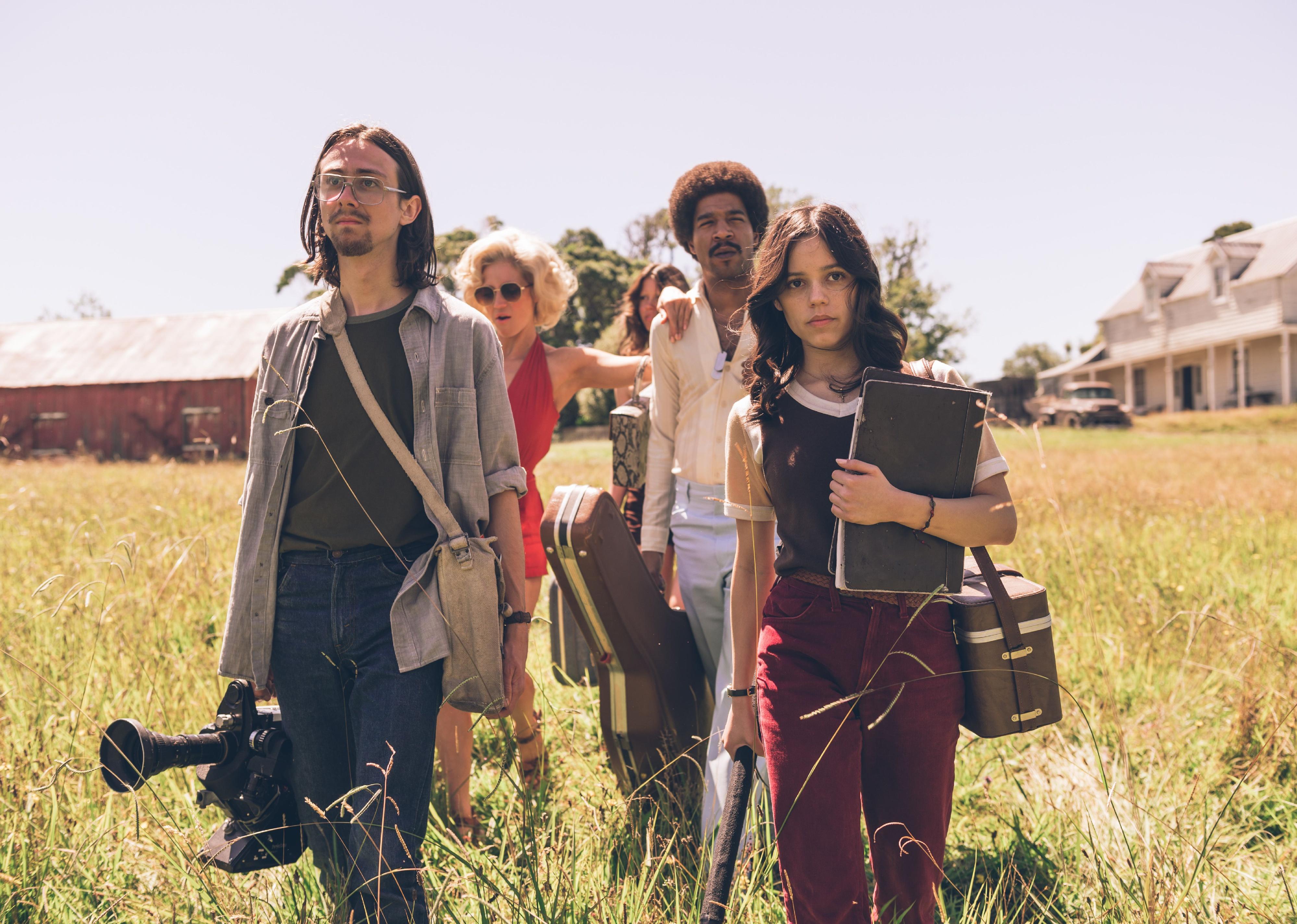 A group of filmmakers and castmembers walk through a field by a red barn carrying film equipment.