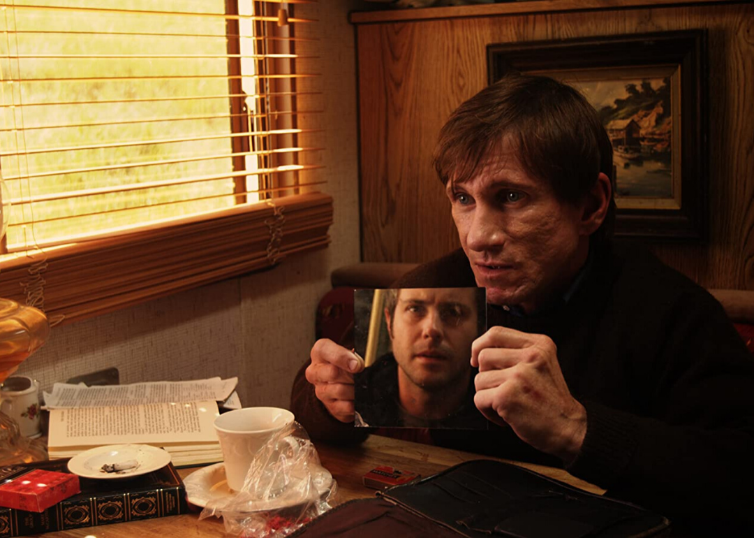A man sits at a small table holding up a mirror to a guy across the table.
