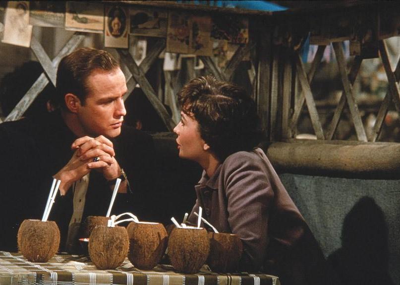 Marlon Brando and Jean Simmons talking together at a table full of coconut shell drinks.