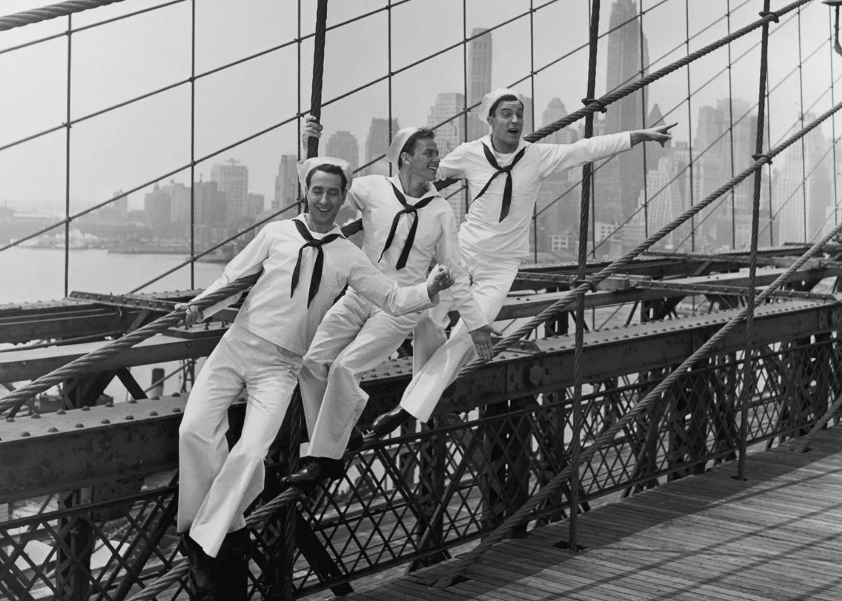 Gene Kelly, Frank Sinatra, and Jules Munshin dressed as sailors dancing and singing on a bridge.