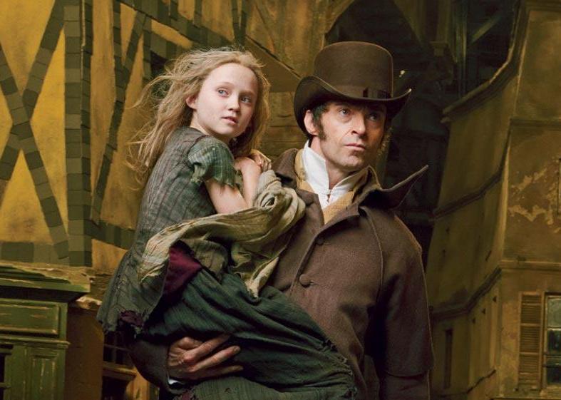 Hugh Jackman in a brown coat and hat carrying a young girl in tattered clothing.