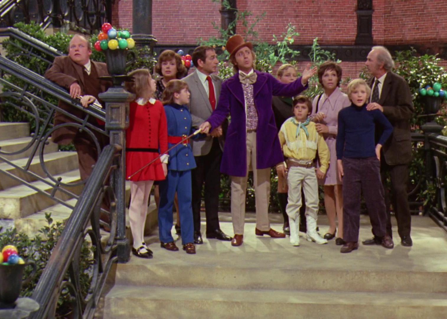 Gene Wilder, as Willy Wonka in a purple coat, showing a group of kids and parents around his factory.