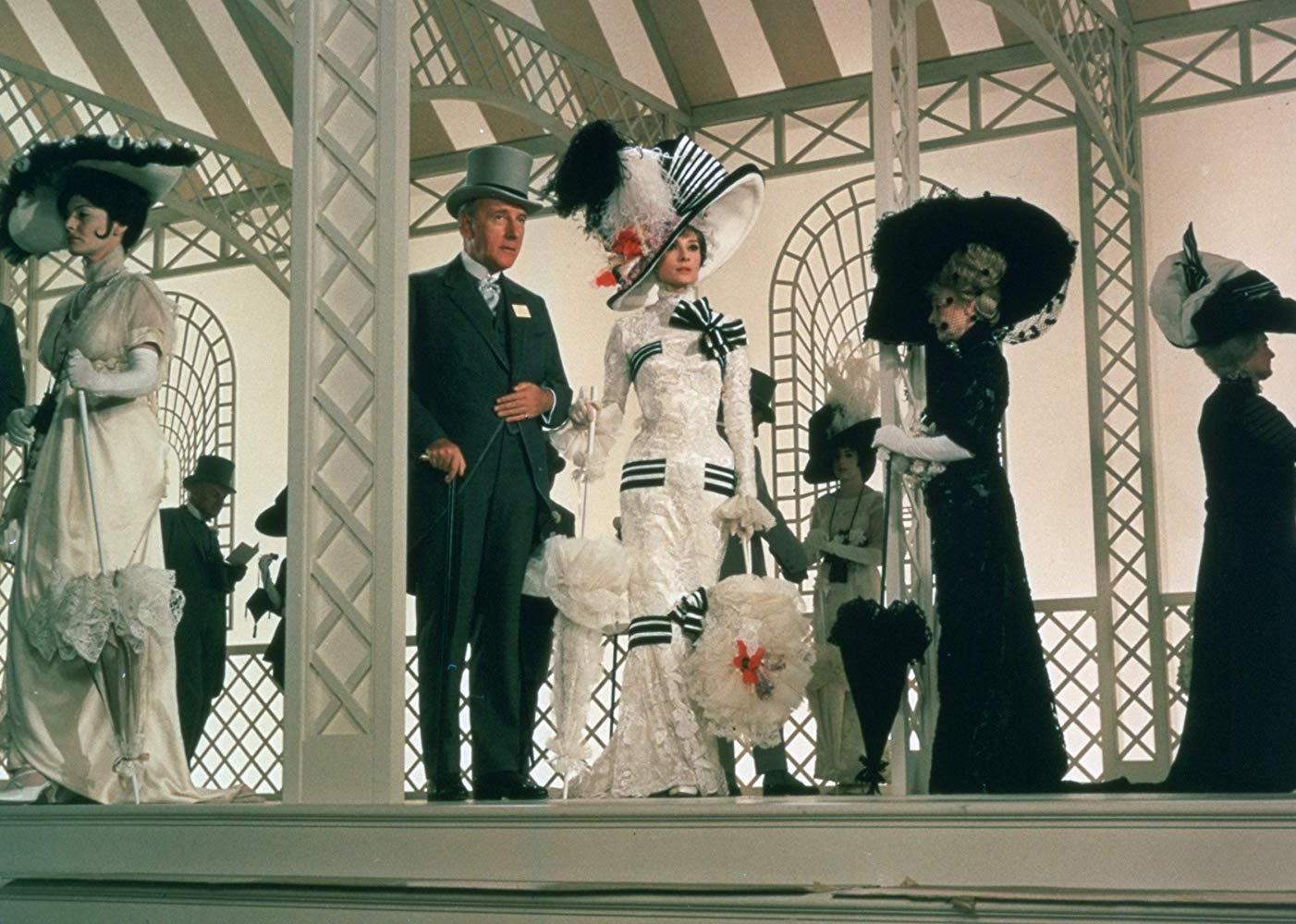 Audrey Hepburn in a beautiful gown and hat arm in arm with a man in a gray suit.