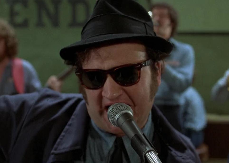 John Belushi in a black hat and sunglasses singing onstage.