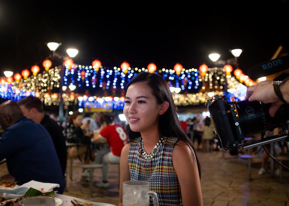 A nicely dressed woman sits at a picnic table with bright lights and other people in the background.