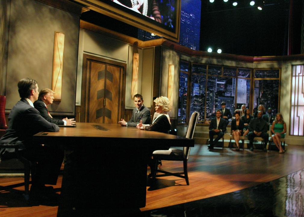 Joan Rivers sits at a table with Donald Trump and the rest of the cast of the show is in the background.