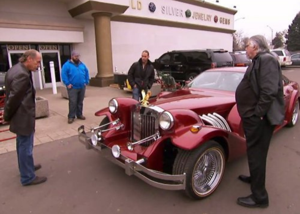 Men walk around a classic car in front of a pawn shop.