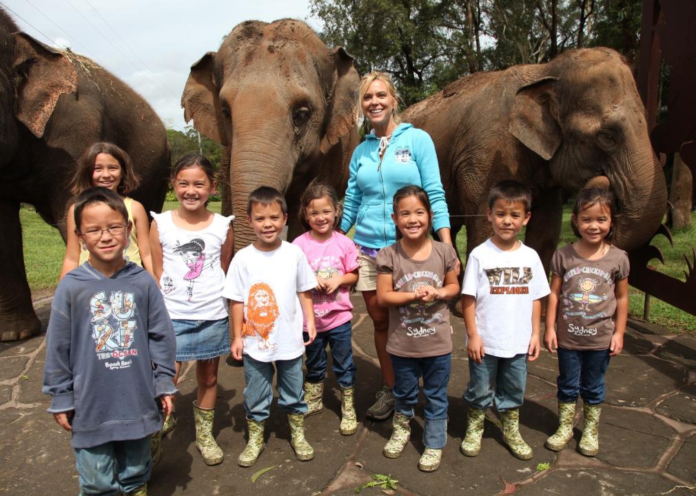 A woman and 8 children stand in front of elephants.