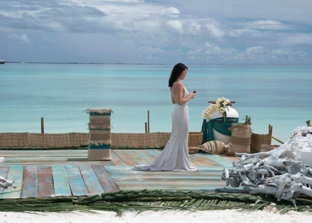 A woman in a long gown holding a rose on a dock in front of turquoise water.