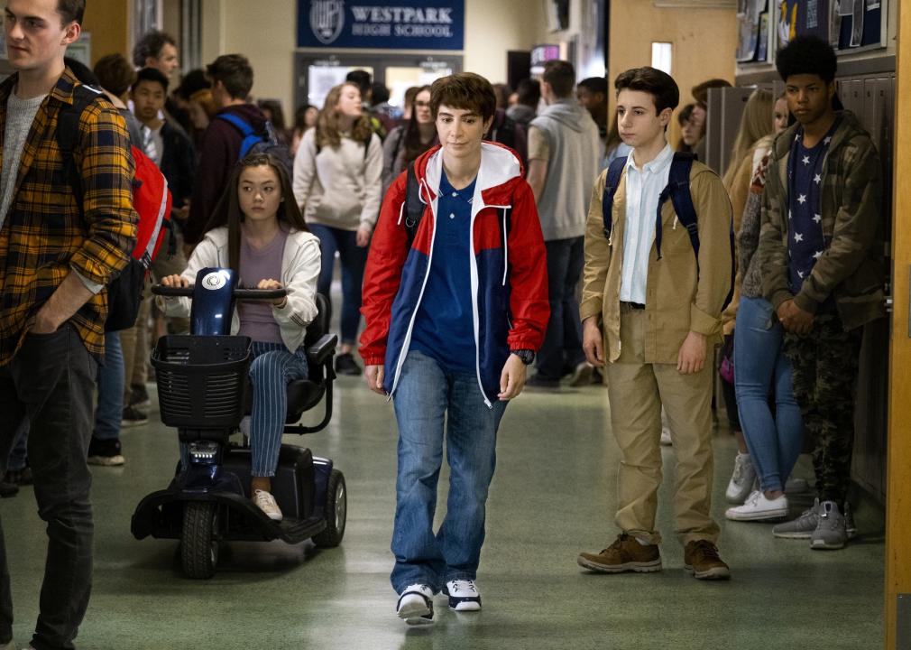 A boy walks down a crowded school hallway next to a girl sitting on a motorized scooter.