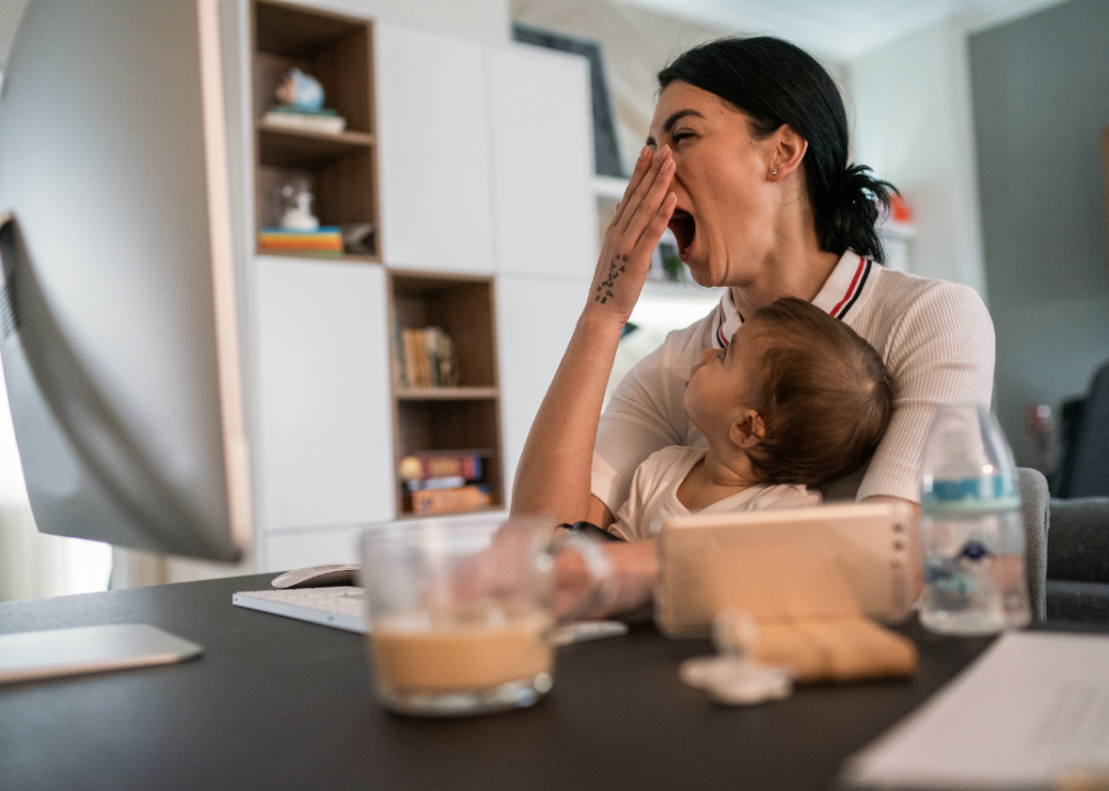 A yawning mother holding a baby while sitting at a computer.