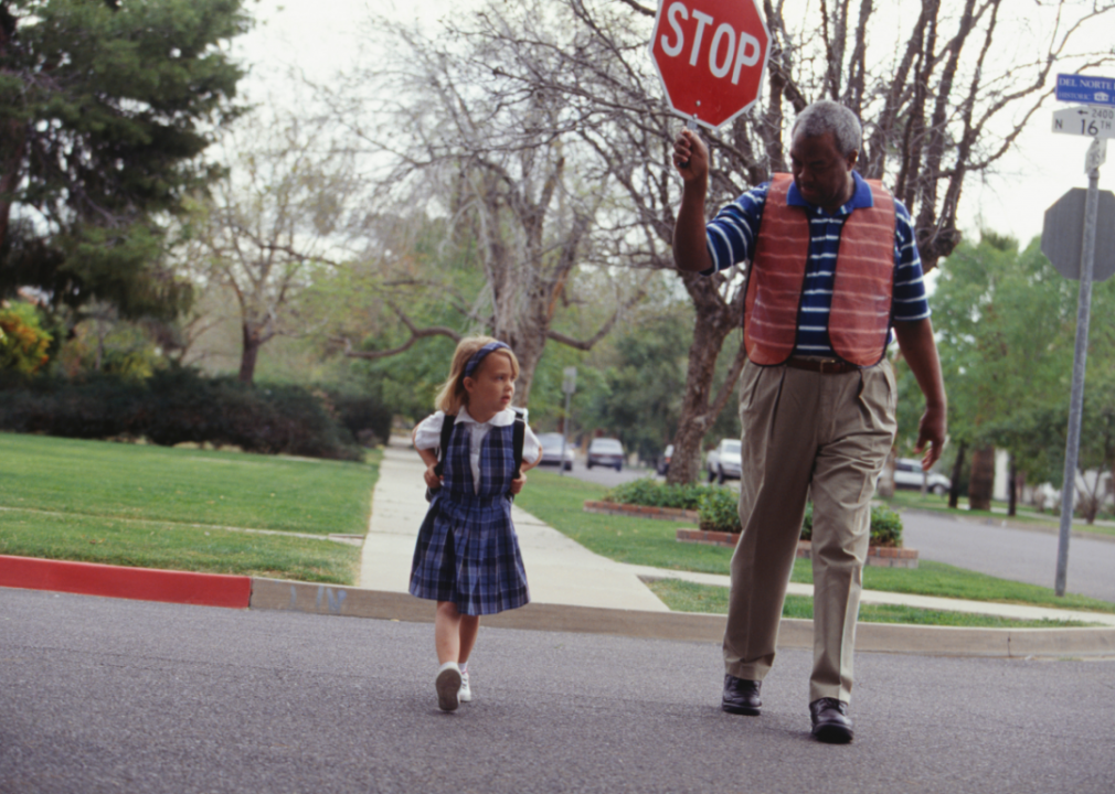 An older man in an orange vest holds up a stop sign as he walks a little girl in a school uniform across the street. 