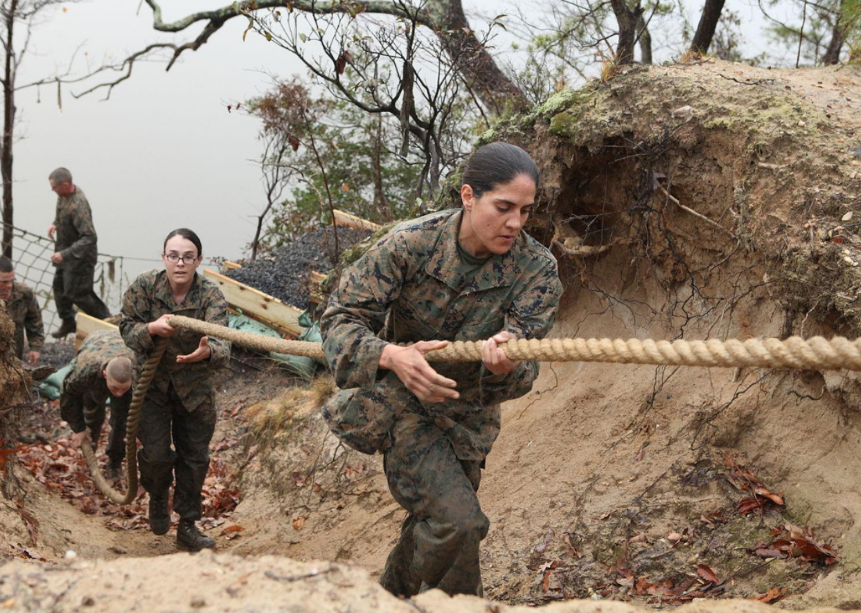 Women in camo walking through mud holding onto a rope.