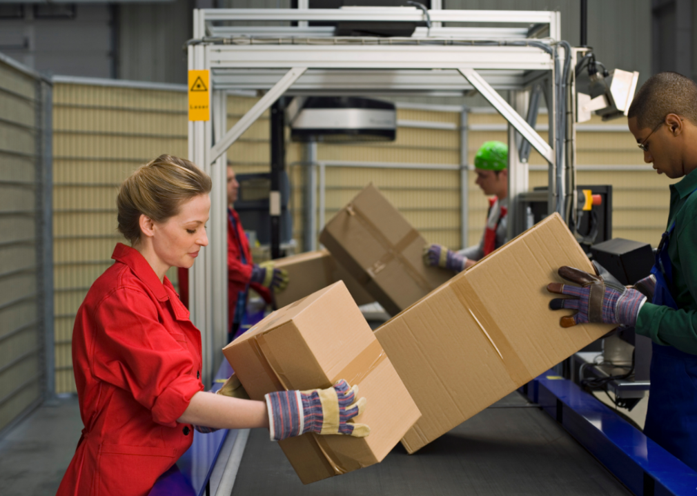 Four people put boxes on a conveyor belt in a warehouse.