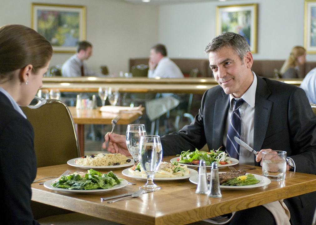 A man in a suit sits across from a young woman at a restaurant while they eat salads.