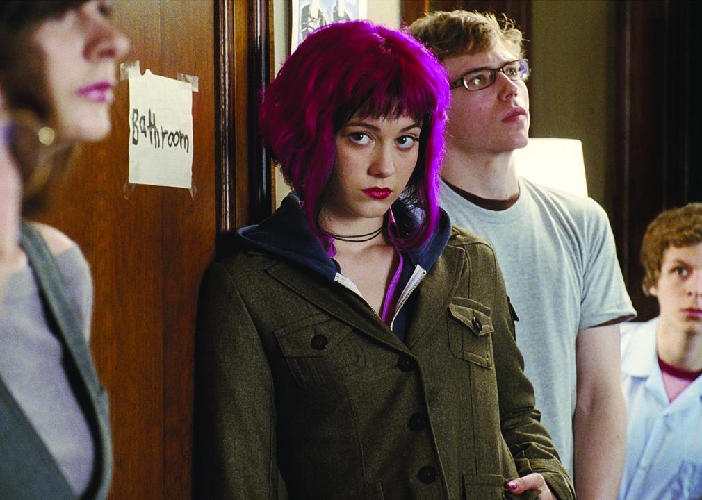 A girl with pink hair waits in a bathroom line while a boy peeks around the corner at her.