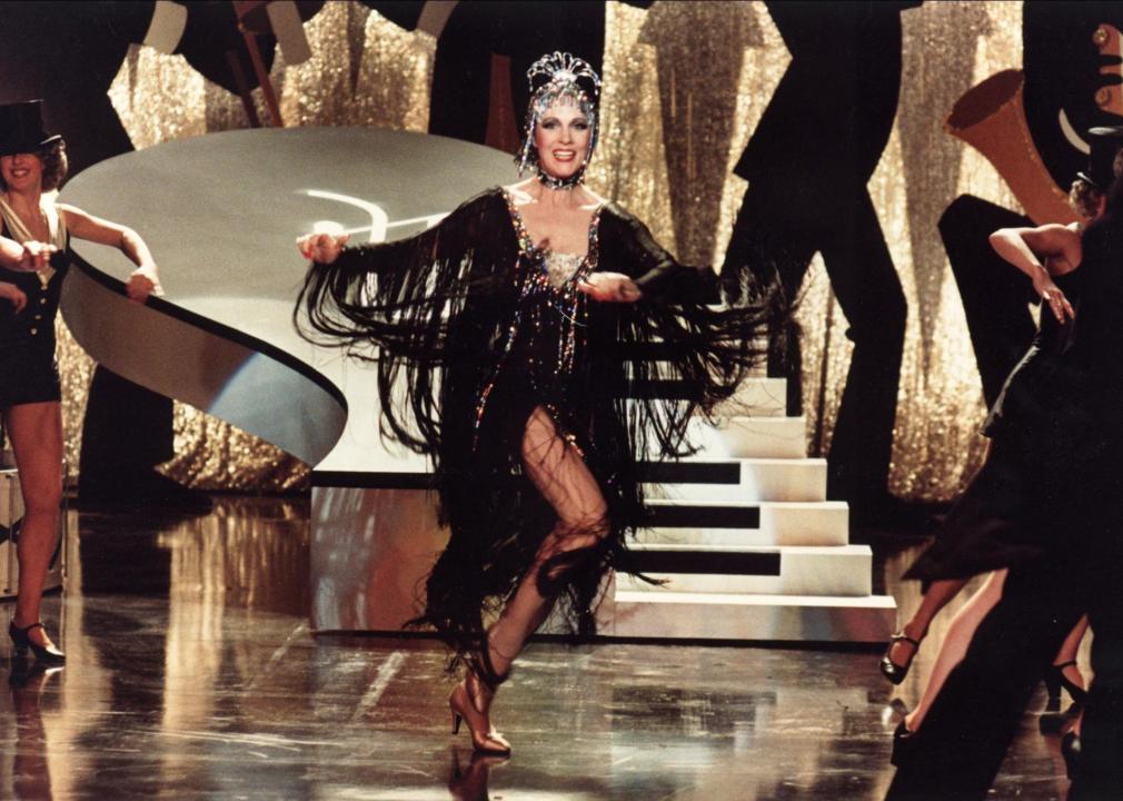 A woman in a long black flapper dress with jewels dances onstage.