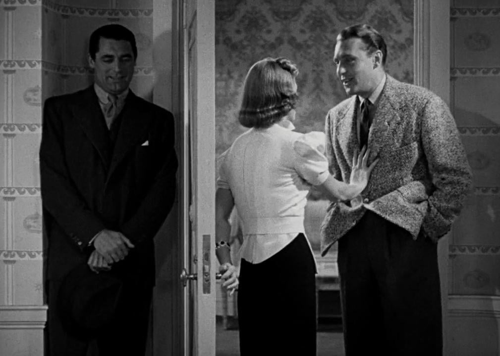 A woman talks with a man at her door while another man hides inside.