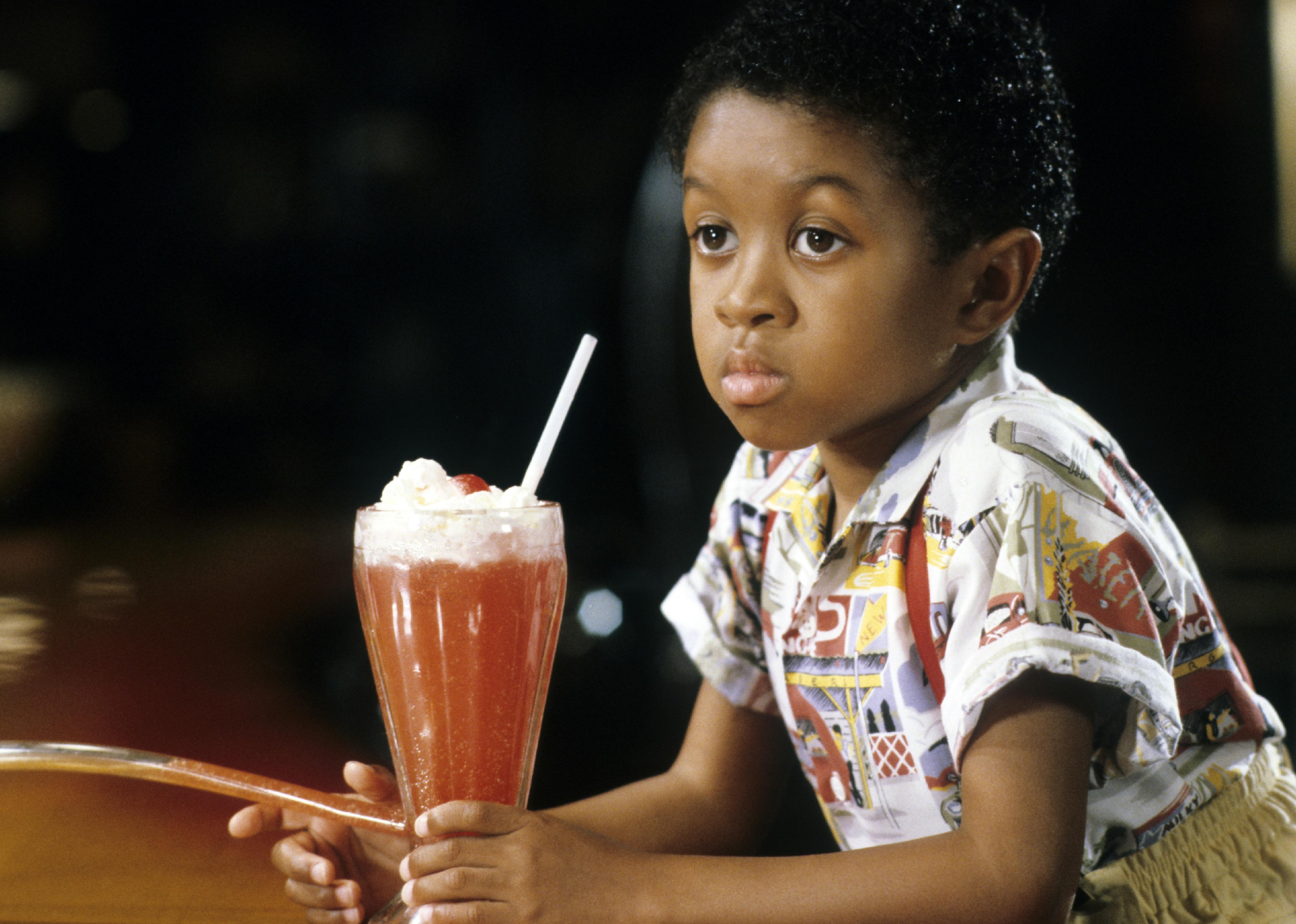 Emmanuel Lewis holding a fruity drink withw whipped cream and a straw.