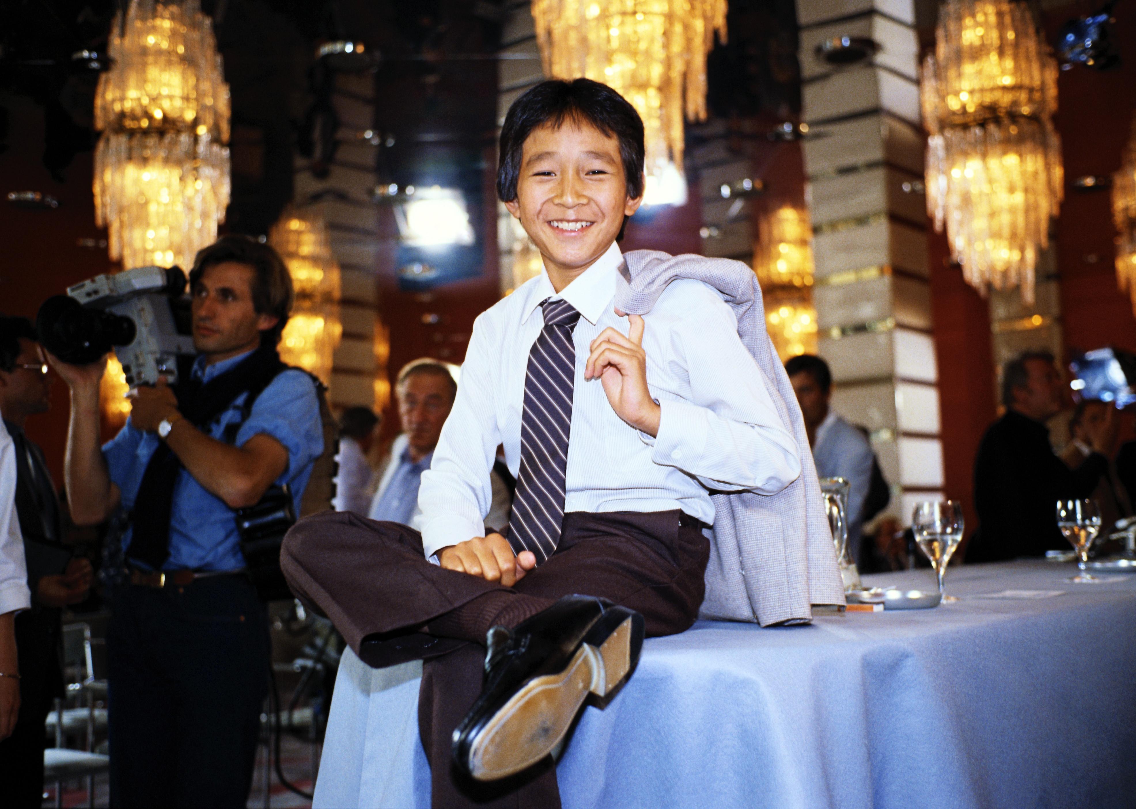 Ke Huy Quan in a suit sitting on a table.