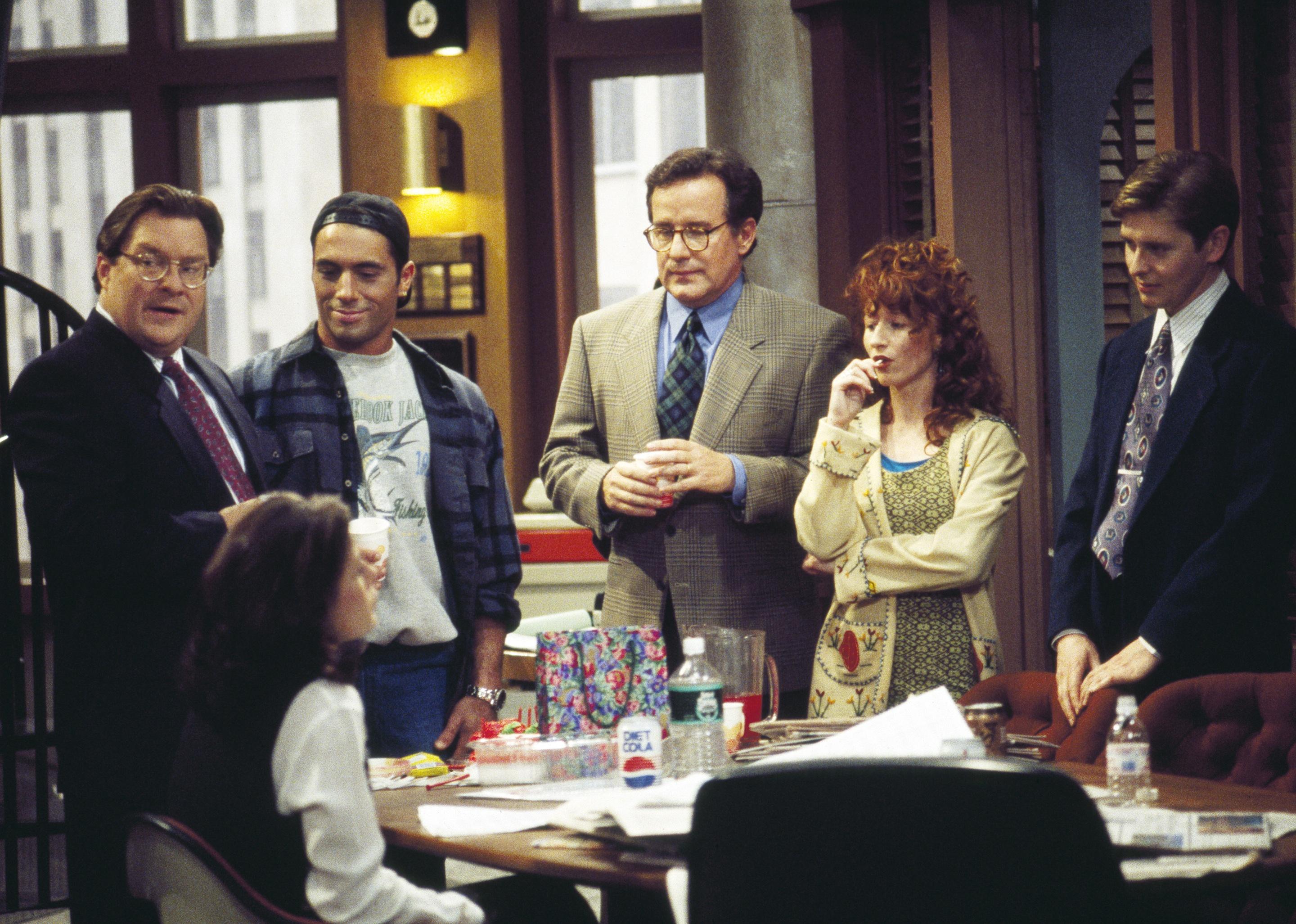 David Foley, Maura Tierney, Phil Hartman, Vicki Lewis, Joe Rogan, and Stephen Root standing around a desk in an office.