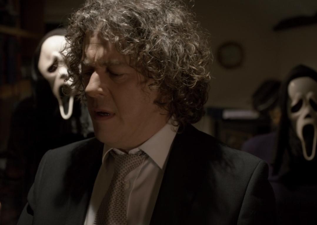 A man in a suit looking on a phone with people in scream masks behind him.