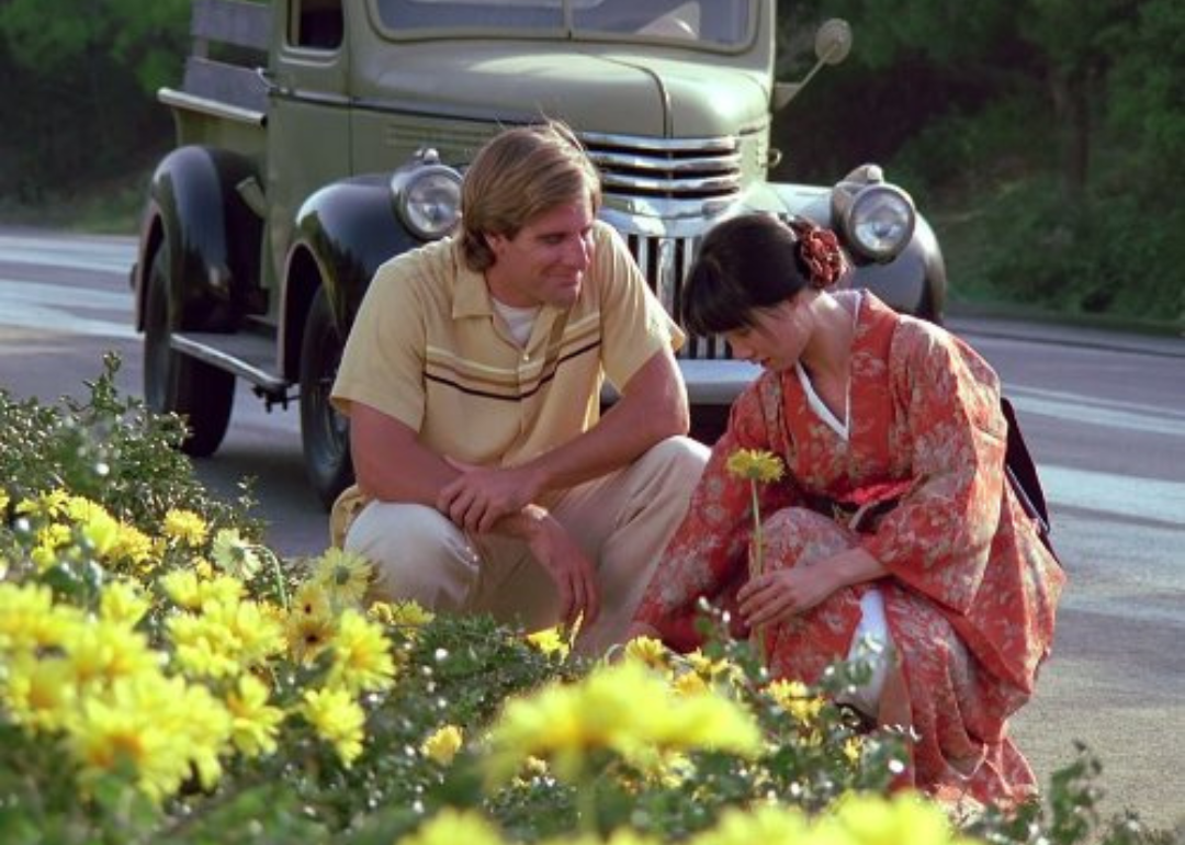 A man stopped on the side of the road in an antique car talking to a woman in a kimono picking yellow flowers.